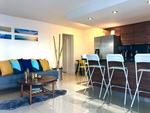 Una sala de estar moderna con sofá, una mesa de centro y sillas. Cuenta con una zona de cocina, un refrigerador y armarios.