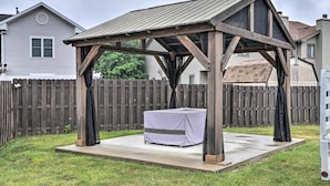Un kiosque en bois avec une table recouverte d’une nappe blanche, installé sur une terrasse en béton dans un jardin, avec une clôture en bois et des maisons en arrière-plan.