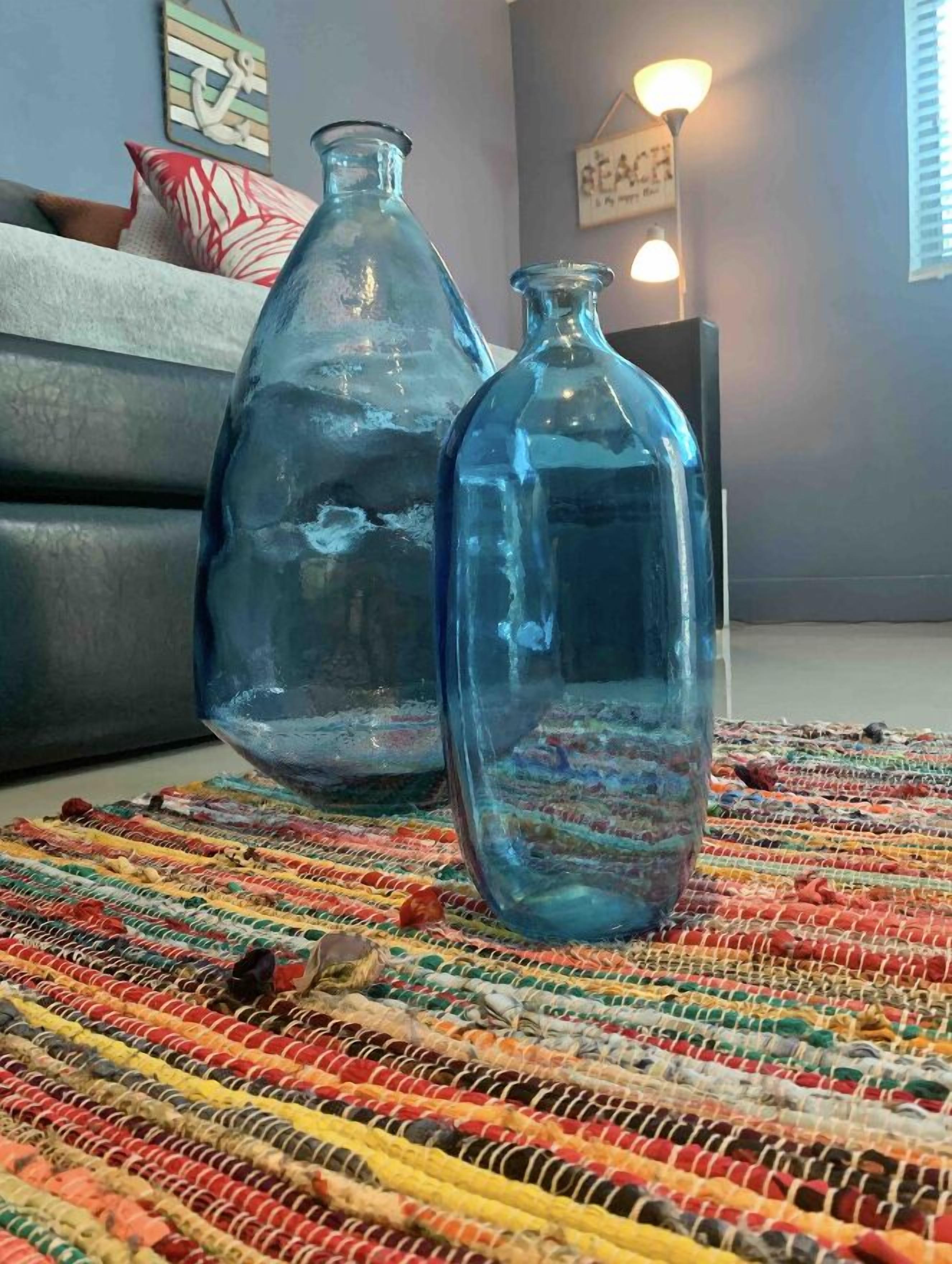 Two clear glass bottles on a colorful woven rug in a living room with a gray couch and a wall-mounted light fixture.