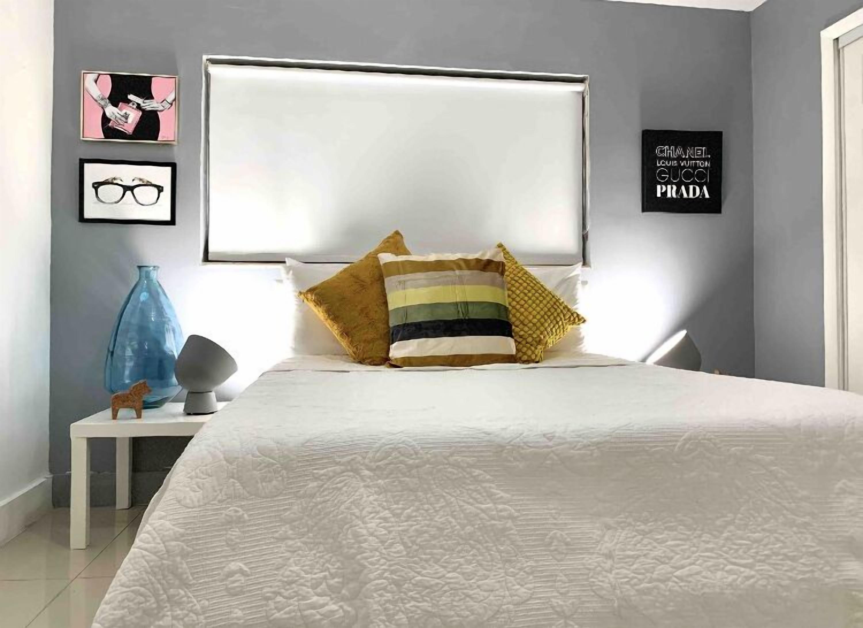 A bedroom with a large bed, a bedside table with a lamp, and a wall with framed pictures and a sign.