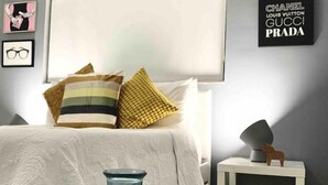 A modern bedroom with a large bed, a bedside table with a lamp, and a wall with luxury brand logos. - THE GROVE 1  10 MIN FROM THE BEACH. In the heart of Coconut Grove. CocoWalk (Miami)