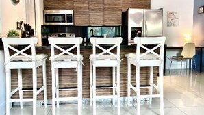 A modern kitchen with wooden cabinets, a microwave, a stove, and a refrigerator. There are three white bar stools in front of the kitchen. - THE GROVE 1  10 MIN FROM THE BEACH. In the heart of Coconut Grove. CocoWalk (Miami)
