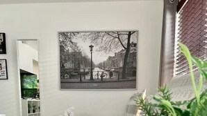 Habitación de hotel con una cama, una mesita de noche con una planta, un espejo, un cuadro en la pared y una ventana con persianas.
