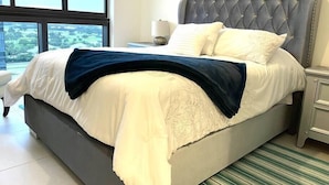 A modern bedroom with a large bed, a grey upholstered headboard, and a view of the city through large windows. - A1608 AWESOME Downtown Doral Apartment (Doral)