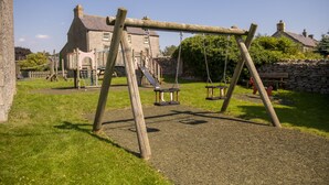 A wooden swing set in a grassy area with a stone wall and a building in the background. - Witton View, Bellerby, Yorkshire Dales (Leyburn)