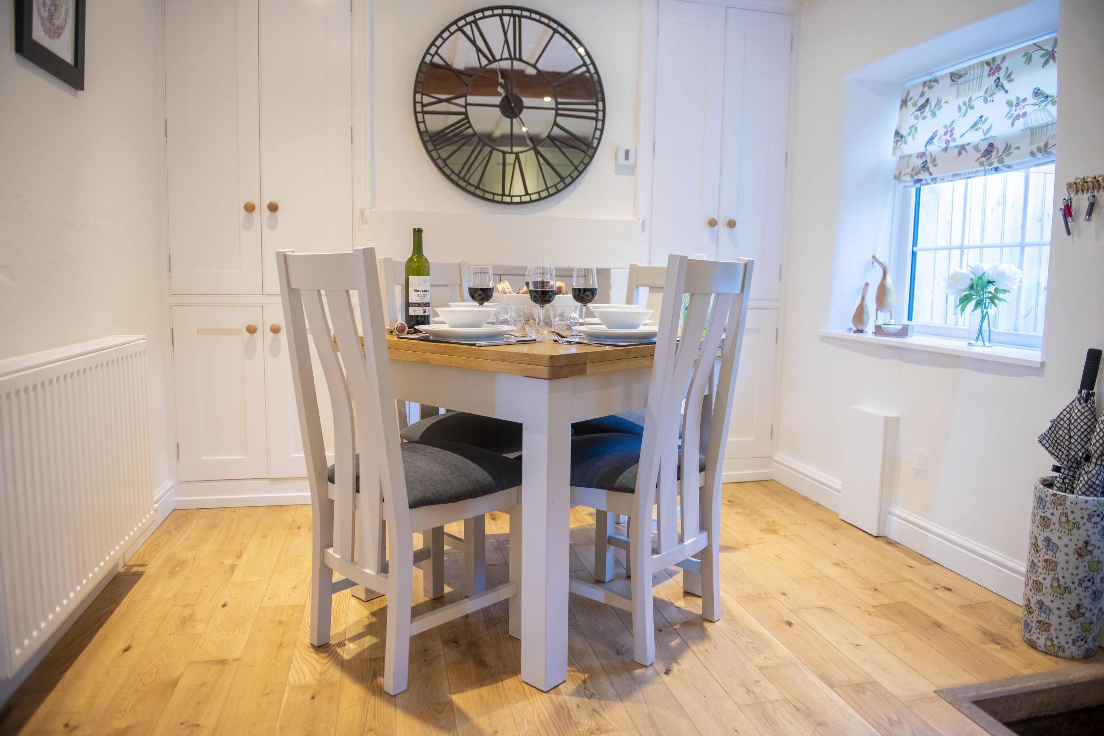 A dining area with a wooden table, white chairs, a large wall clock, and a window with a view of the outside.