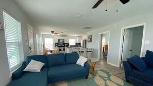Un salon moderne avec un canapé bleu, un coin repas avec des chaises, une cuisine équipée et un ventilateur de plafond.