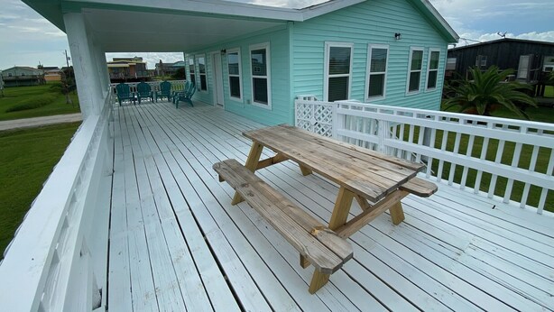 Une terrasse couverte avec une table et des chaises en bois, une balustrade blanche et, au fond, une maison bleu clair.