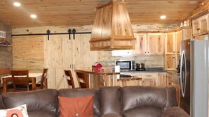 Una sala de estar acogedora con un sofá de cuero marrón, paredes de madera, un ventilador de techo y una zona de cocina con refrigerador y microondas.