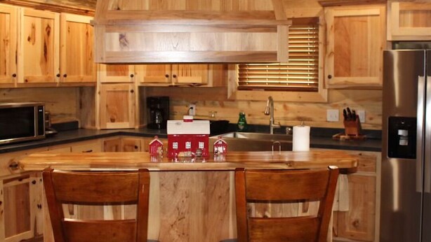Cocina de estilo cabaña de madera con una isla central, dos sillas de madera, un refrigerador de acero inoxidable y armarios de madera.