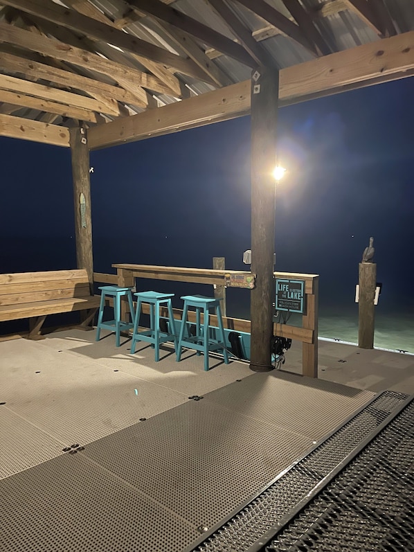 Un espace extérieur abrité, avec des bancs en bois et des tabourets bleus, un panneau indicateur portant un message, et une vue sur l’eau.