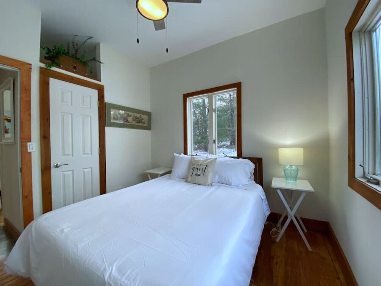 Un dormitorio con una cama grande, una mesita de noche con lámpara, una ventana con vistas a árboles y una puerta con marco de madera.