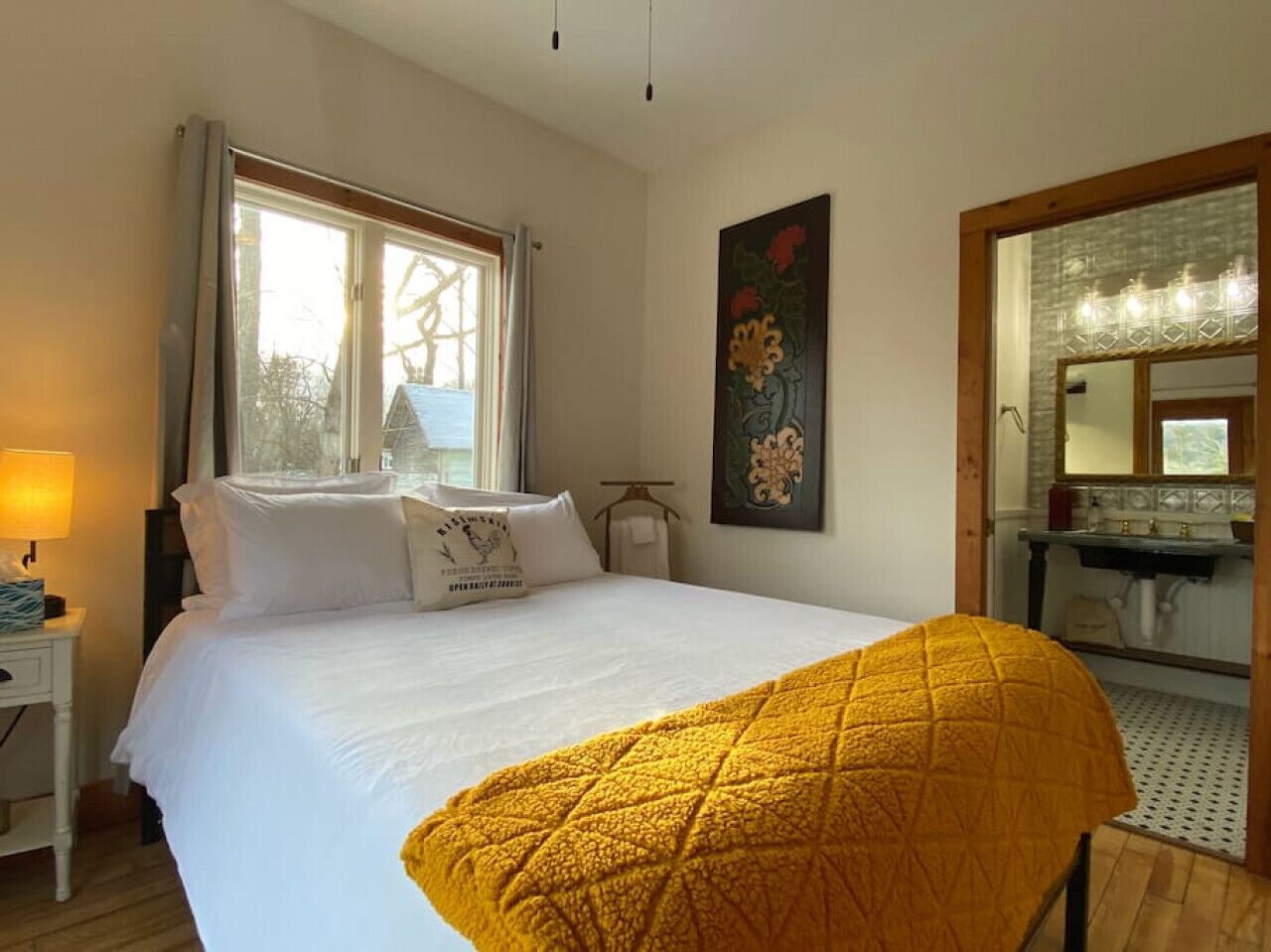Une chambre d’hôtel avec un grand lit, une table de chevet avec une lampe, une fenêtre avec des rideaux, un tableau au mur et une salle de bain visible à travers une porte ouverte.