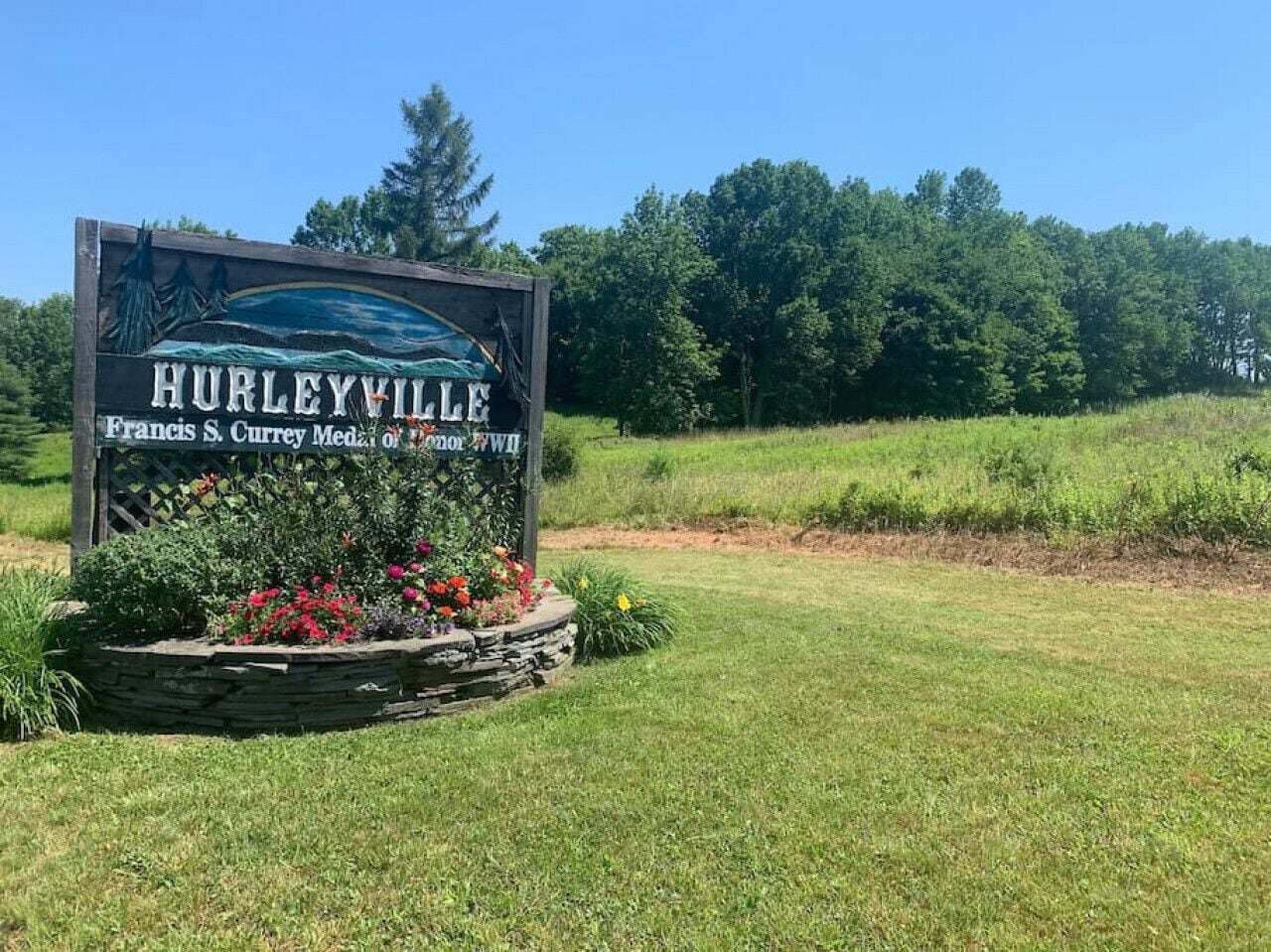 A sign for Hurleyville with a scenic landscape and a stone planter with flowers.
