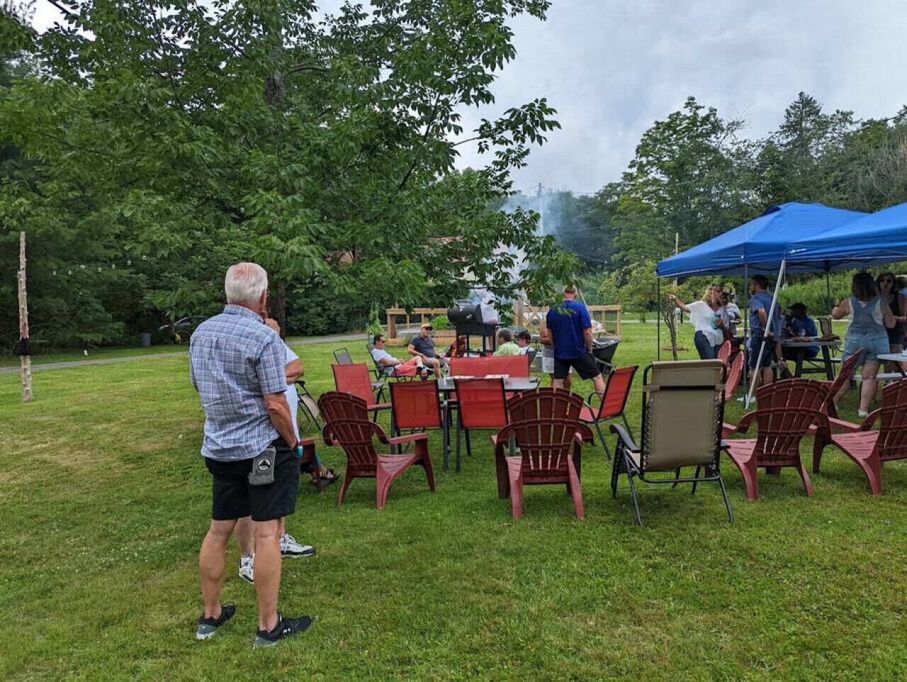 An outdoor gathering with people seated at tables under a blue canopy, surrounded by grass and trees.