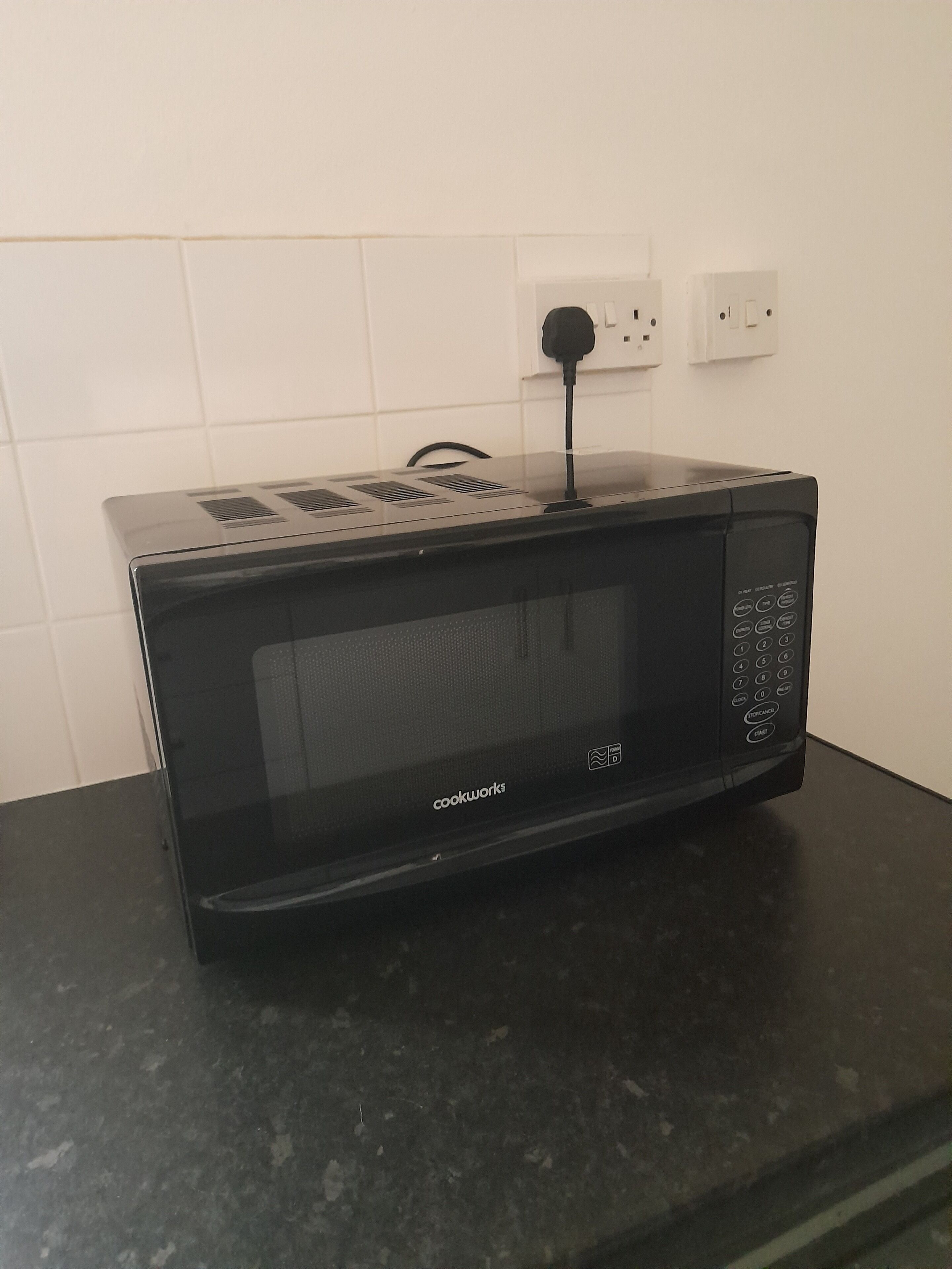 A black Cookware microwave on a dark countertop with a power outlet in the background.