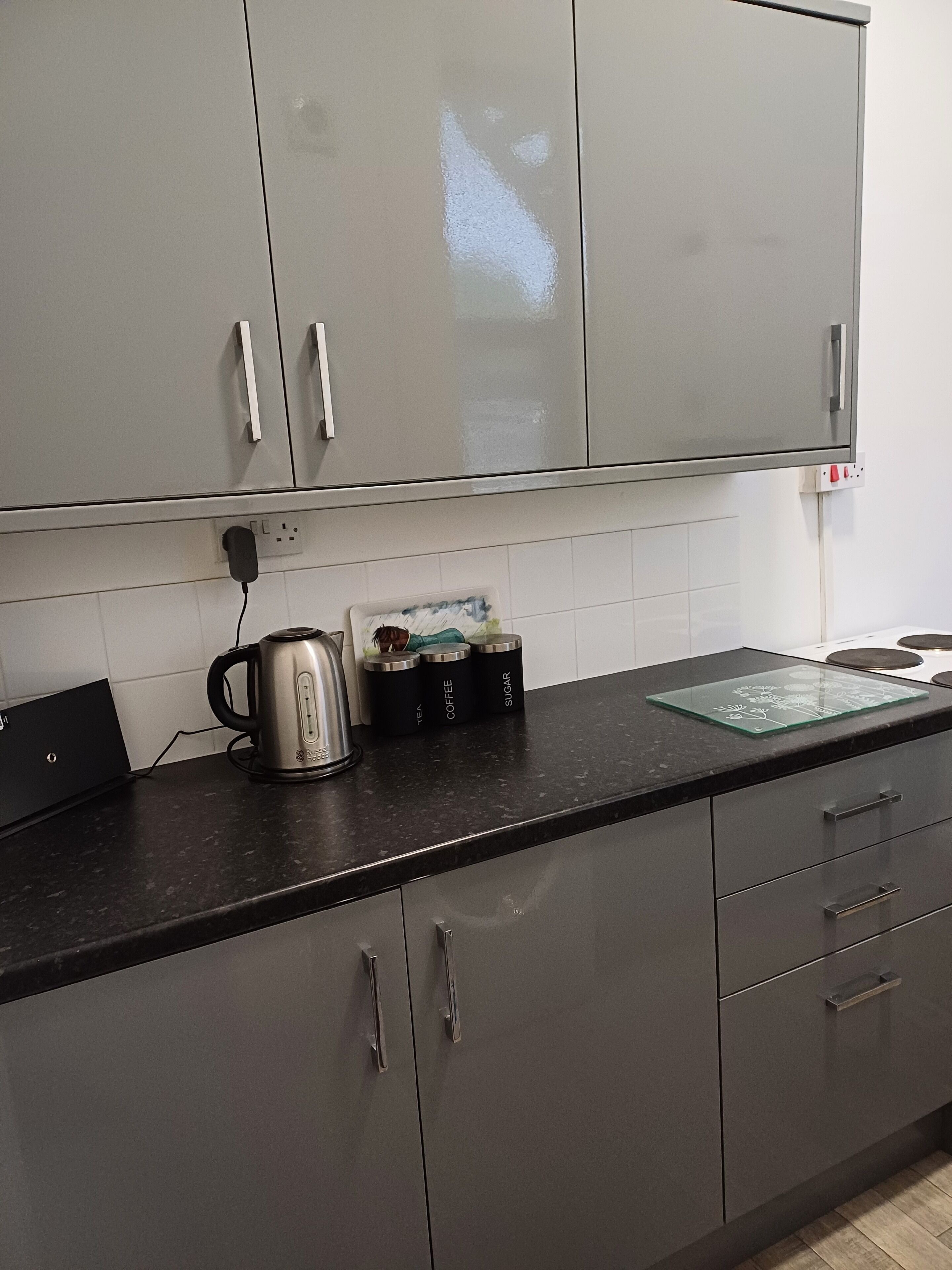 Una cocina con armarios grises, encimera negra y un hervidor. Hay tres recipientes etiquetados como té, café y azúcar.