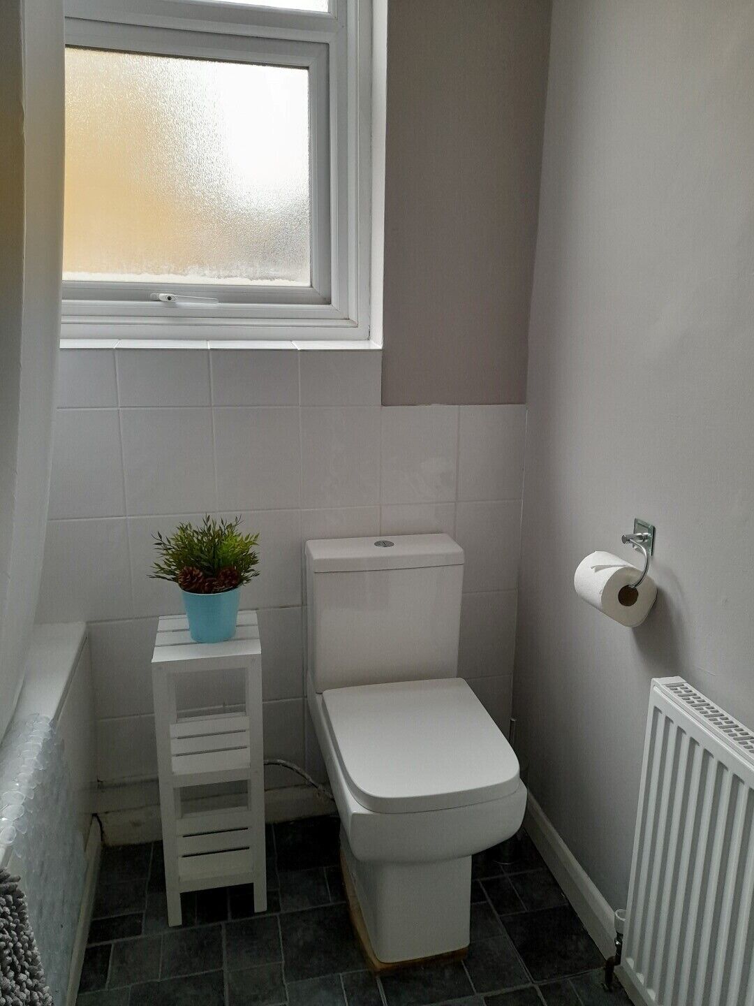 Een kleine badkamer met toilet, een raam, een radiator en een klein krukje met een plant.