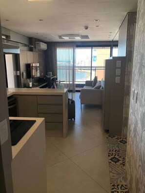 Quarto de hotel com cozinha compacta, área de estar com sofá e pequena mesa, janela ampla com vista para a praia e tapete estampado.