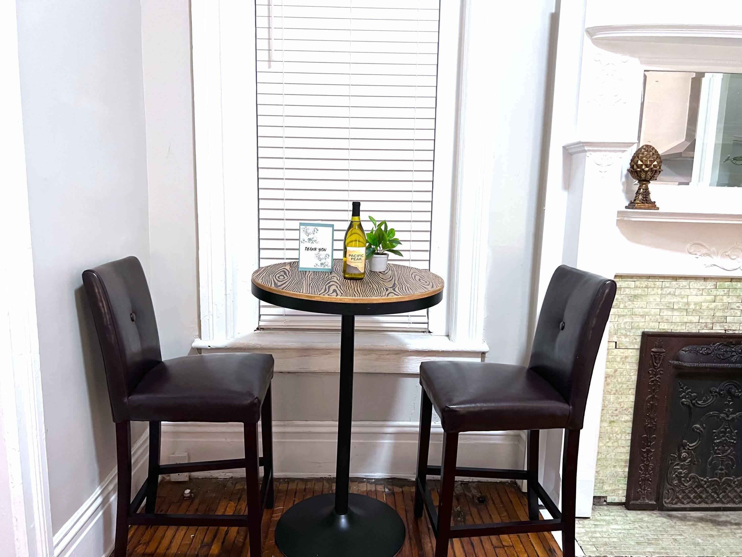 Une pièce d’angle avec une table ronde, deux chaises, une fenêtre avec des stores, une cheminée et une petite plante posée sur la table.