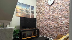 Un salon avec un téléviseur à écran plat posé sur un meuble en bois, un mur décoré d’un motif en briques, une horloge et un canapé marron.