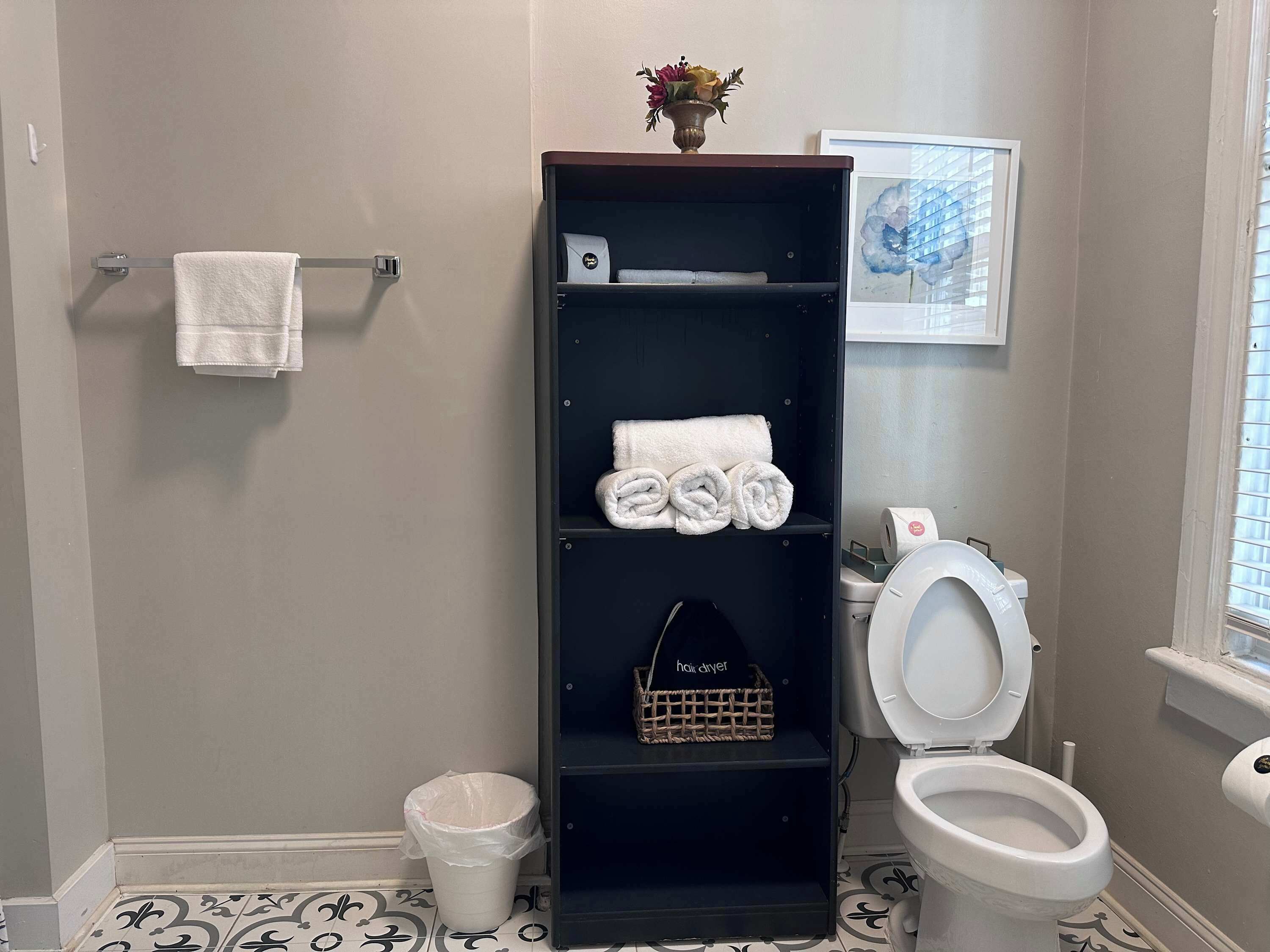 Un baño con inodoro, toallero, repisa con toallas, cubo de basura y un cuadro en la pared.