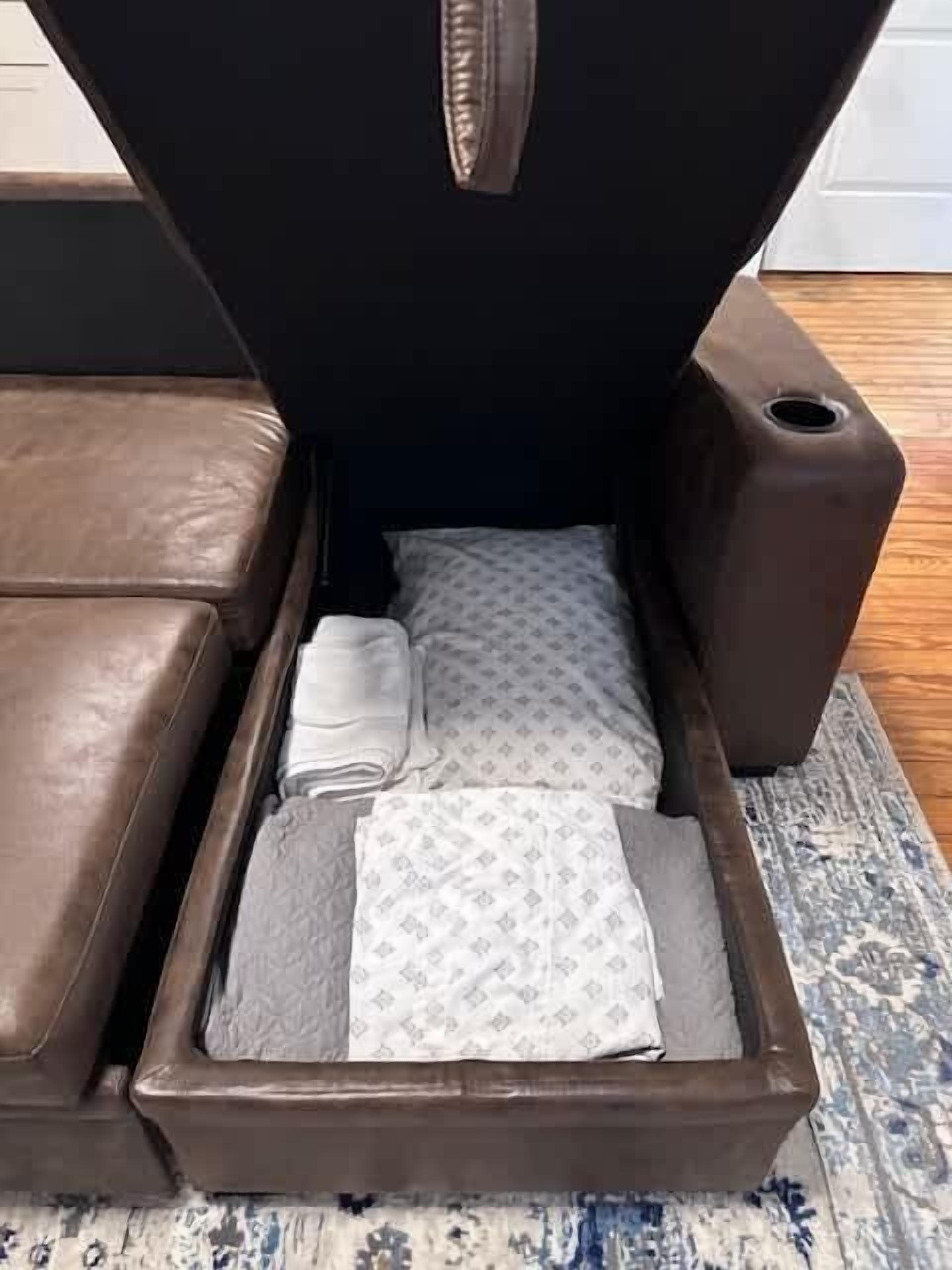 Ein braunes Ledersofa mit Stauraum darunter. Im Fach liegen gefaltete, weiß-graue Decken.
