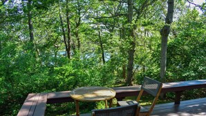 A wooden deck with a round glass table and two wicker chairs, surrounded by lush greenery and a body of water in the background. - Four bedroom home within walking distance to Bayside Brewster beaches! (Brewster)