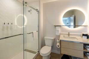 A modern bathroom with a glass shower enclosure, a toilet, a sink with a mirror, and toiletries on the counter. - Towneplace Suites by Marriott Canton Riverstone Parkway (Canton)