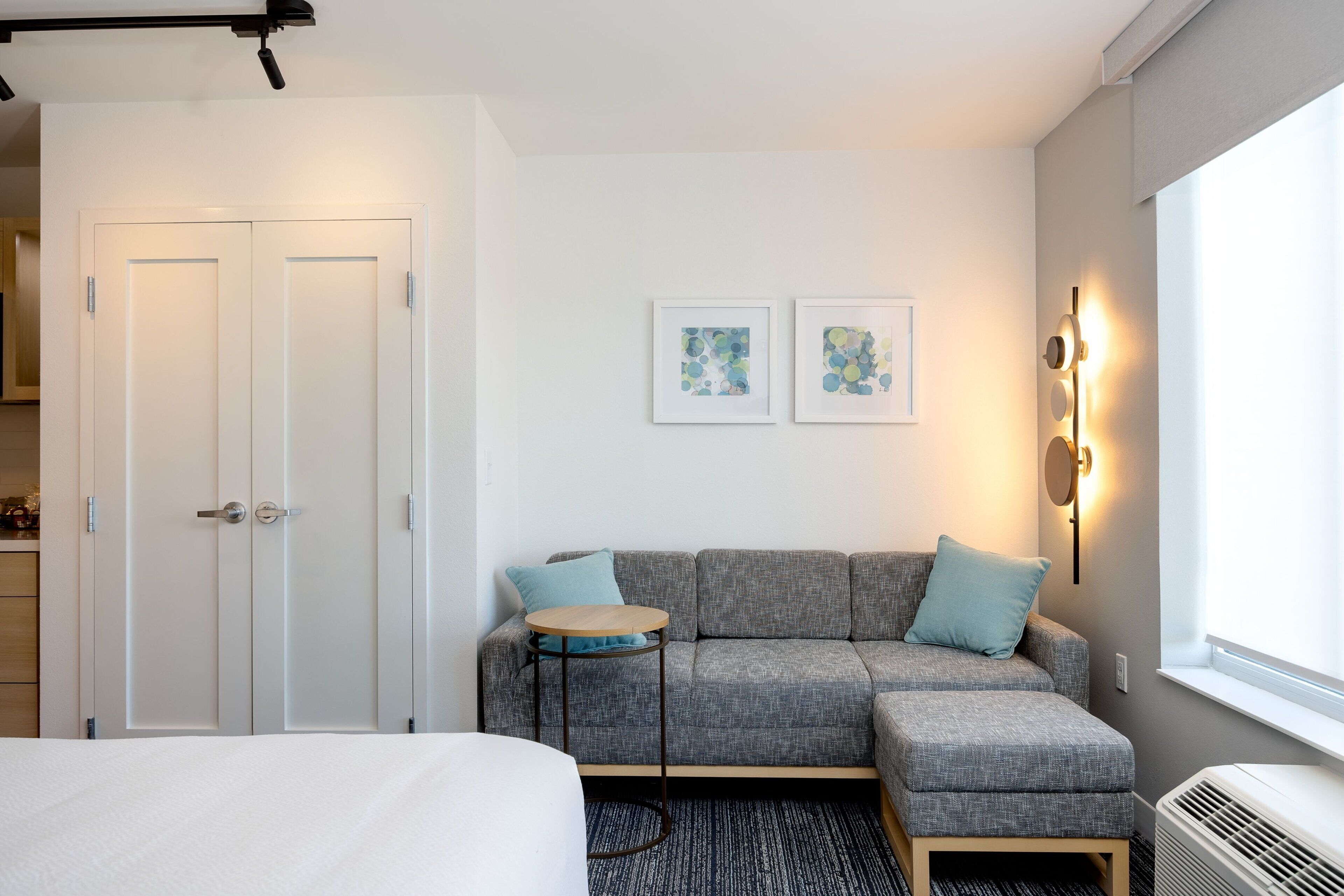 Una habitación de hotel moderna con un sofá gris, una pequeña mesa redonda, una cama y dos cuadros en la pared.