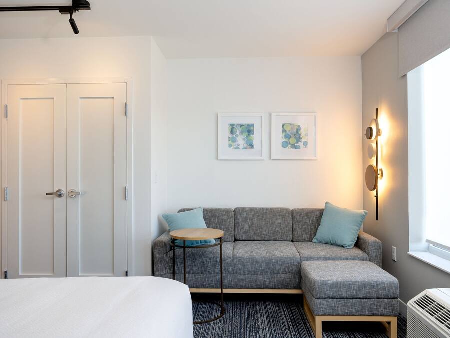 Una habitación de hotel moderna con un sofá gris, una pequeña mesa redonda, una cama y dos cuadros en la pared.