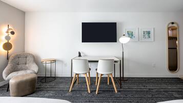A modern hotel room with a grey armchair, a small round table, a desk with two chairs, a flat-screen TV, and a mirror on the wall.