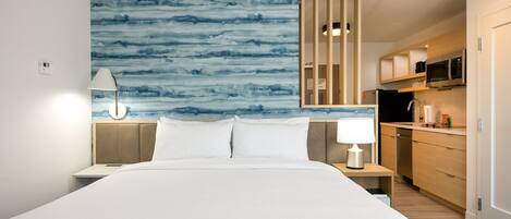Una habitación de hotel moderna con una cama grande, una mesita de noche, un escritorio, una kitchenette y una pared azul con motivos abstractos.