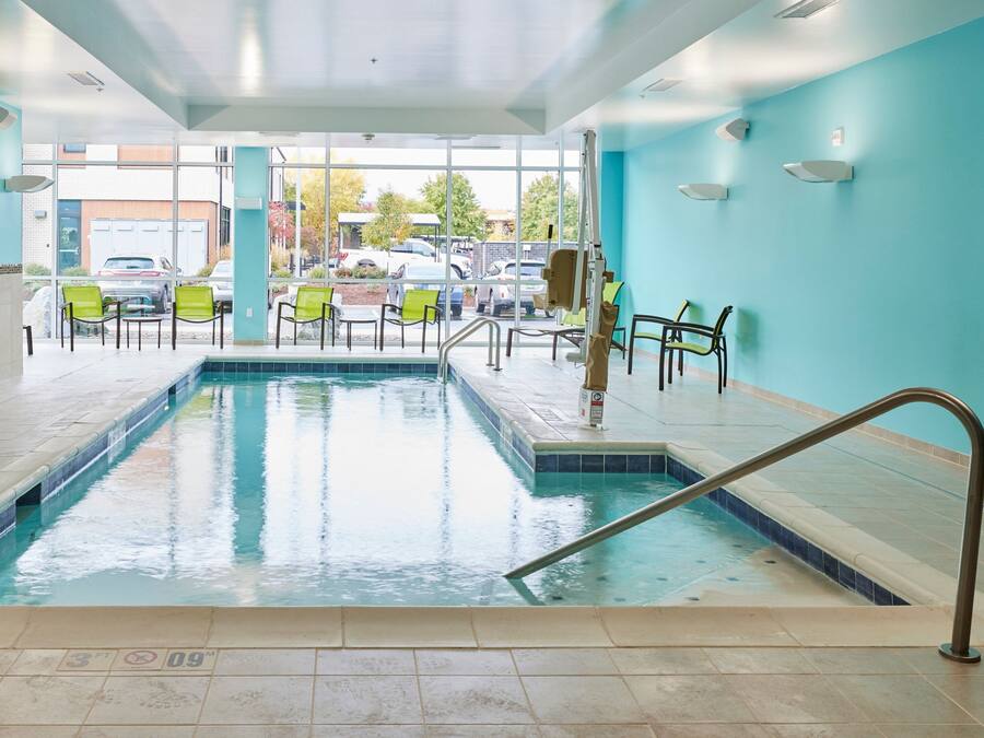 En indendørs swimmingpool med klart, blåt vand, omgivet af grønne stole og flisegulv.