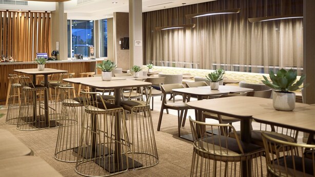 Un hall d’hôtel moderne doté de plusieurs espaces de détente, notamment des tables et des chaises rondes, ainsi qu’un bar avec des tabourets.