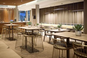 Un moderno vestíbulo de hotel con varias zonas de estar, incluidas mesas y sillas redondas, y un área de bar con taburetes.