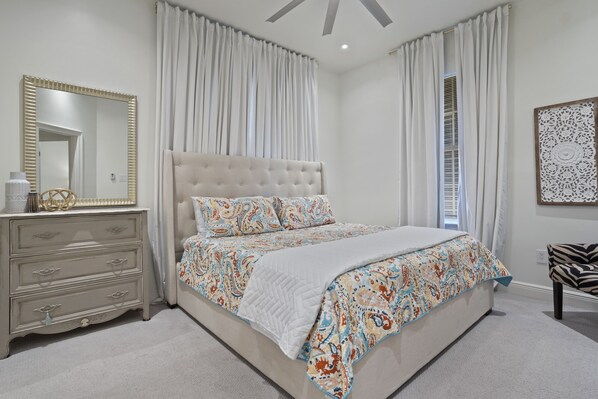 A bedroom with a tufted headboard, a patterned bedspread, a bedside table with a lamp, and a large mirror. - BSL Dream Baycation - 2 blocks to Old Town! (Bay St. Louis)
