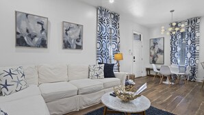 Un salon moderne avec un canapé blanc, des tableaux abstraits, des rideaux à motifs et un coin repas comprenant une table et des chaises.