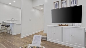 Un salon moderne doté d’un téléviseur à écran plat, de meubles blancs et d’un coin repas avec des tabourets de bar.
