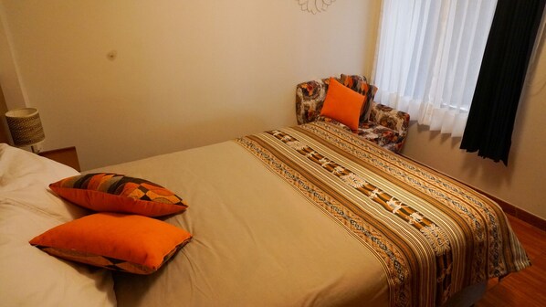 A bedroom with a bed, two pillows, a patterned blanket, a lamp, and a window with curtains.