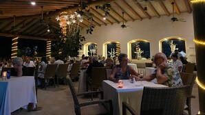 An indoor dining area with wooden beams and hanging plants. Tables are set with white tablecloths and candles. - Luxurious Pacifica Grand Senior Suite (Zihuatanejo)