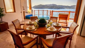 A dining area with a round table set for six, surrounded by wooden chairs. A large window offers a view of a body of water and distant hills. - Luxurious Pacifica Grand Senior Suite (Zihuatanejo)