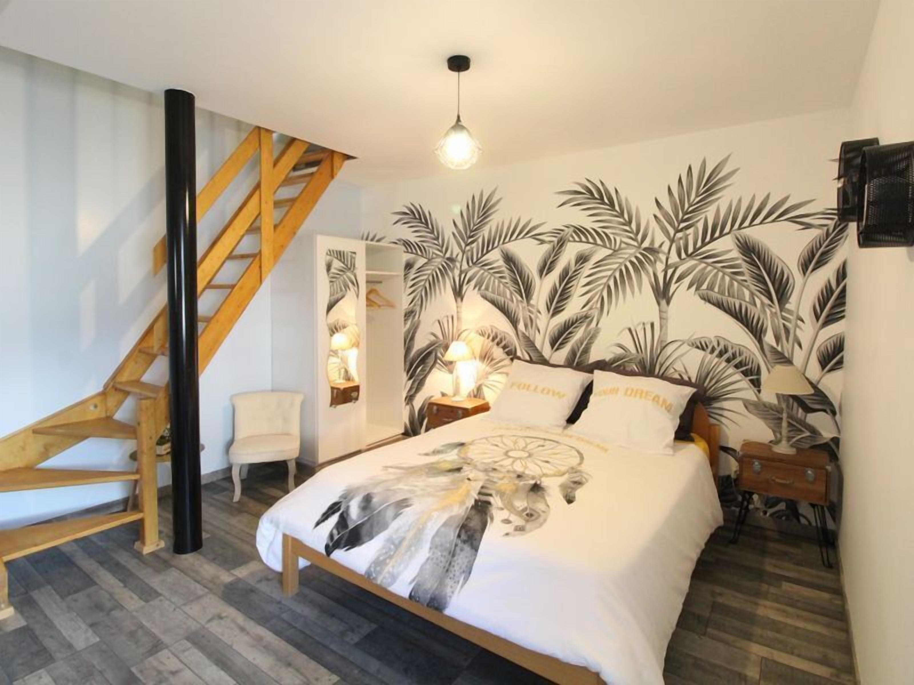 A bedroom with a bed, a nightstand, a lamp, a chair, a wooden staircase, and a wall with a leaf pattern.