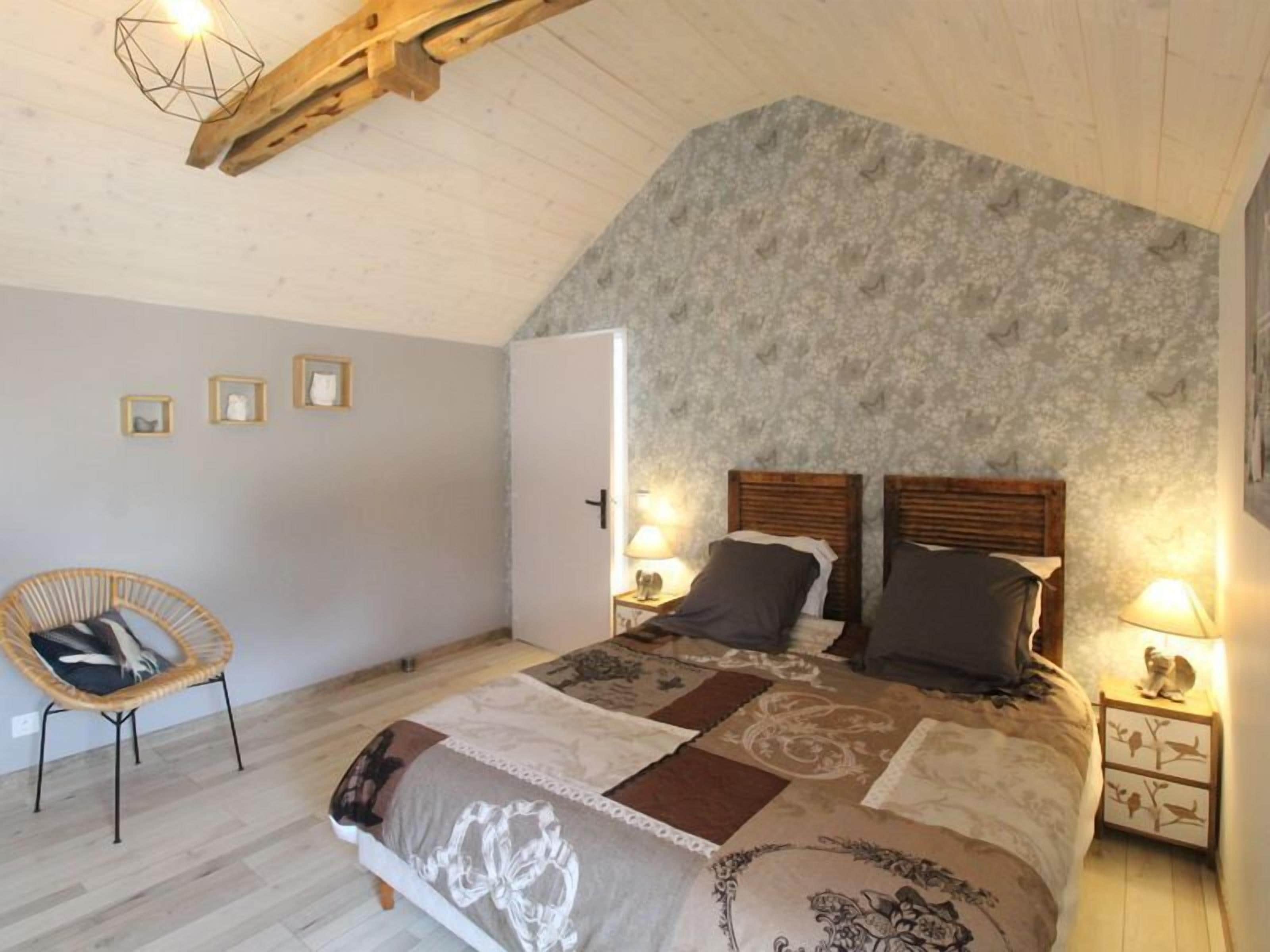 Un dormitorio con techo inclinado, cama con cubrecama estampado, dos lámparas de noche, una silla de mimbre y una puerta que da a otra habitación.