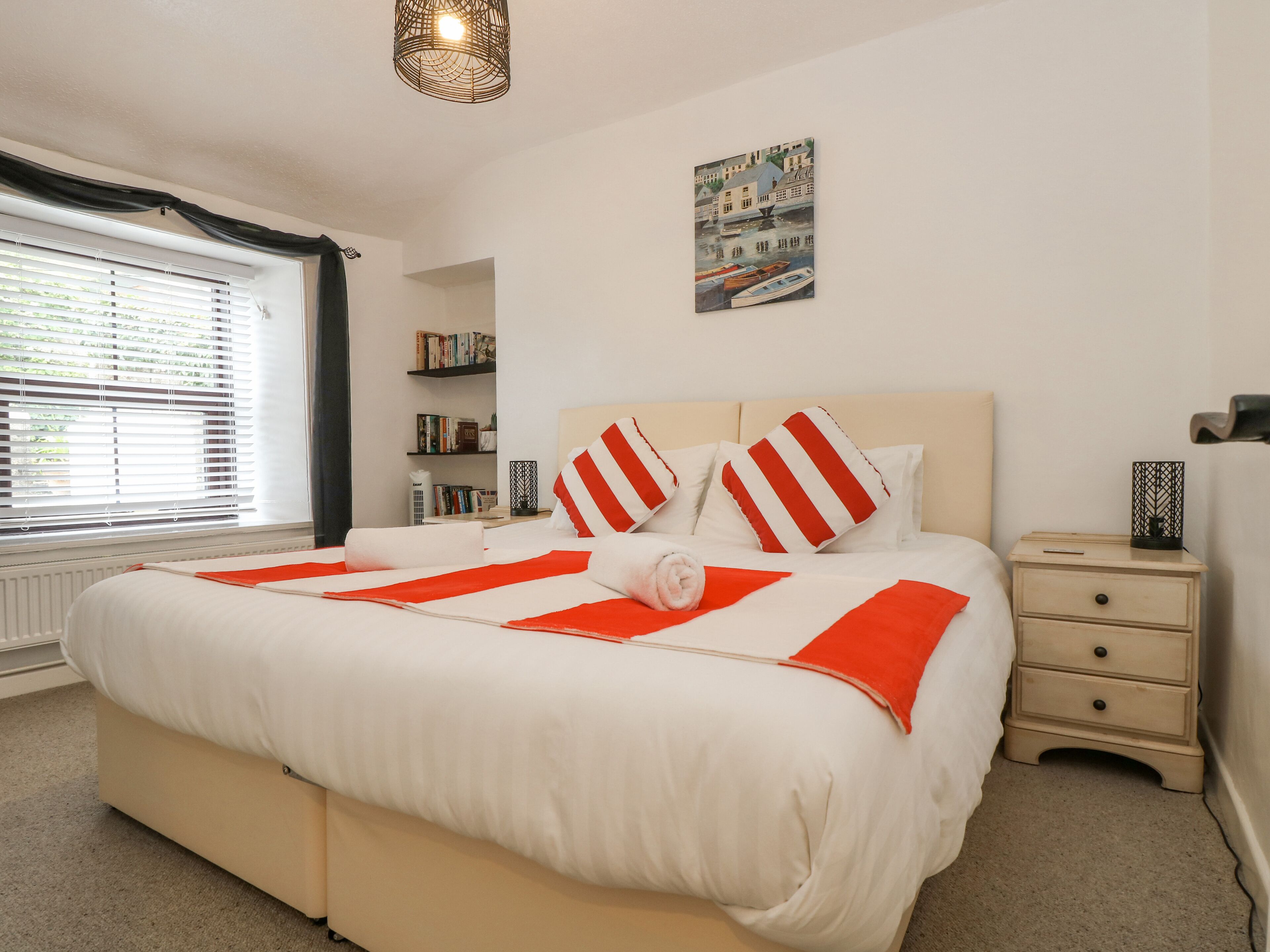 A neatly made bed with red and white striped pillows, a bedside table with a lamp, and a bookshelf with books.