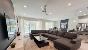 Een moderne woonkamer met een hoekbank, een flatscreen televisie en een eethoek, met een keuken op de achtergrond.