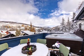 Un patio cubierto de nieve con un jacuzzi, una mesa con sillas y una vista panorámica de un pueblo nevado y montañas.
