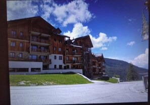 Ein mehrstöckiges Holzgebäude mit Balkonen, einer Grünfläche davor und einer Bergkulisse im Hintergrund.
