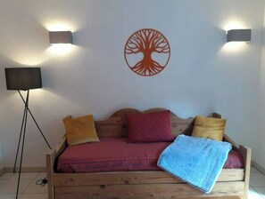A room with a wooden bed, red and yellow pillows, a blue blanket, and a wall-mounted tree decal. There is a floor lamp and a TV on a stand.
