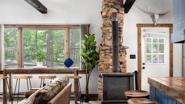 Una sala de estar acogedora con chimenea de piedra, vigas de madera, un gran ventanal con vistas a un bosque y una zona de bar rústica.